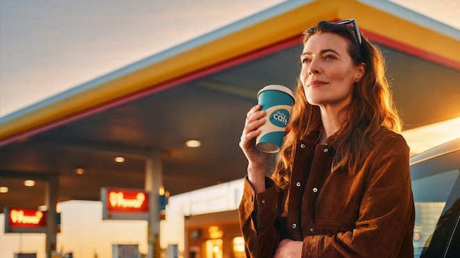copy for the image is: A smiling woman wearing a brown suede jacket stands next to a car at a Shell gas station during golden hour. She is looking peacefully into the distance while holding a blue and white Shell Café paper coffee cup.