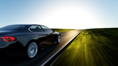 A black car speeding on an asphalt road with motion blur effect on the surrounding grass and sky, indicating high speed, with a bright sun near the horizon in the background.