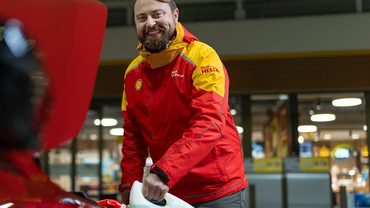 Usměvavá venkovní obsluha Shell pomáhá zákazníkovi s autem.