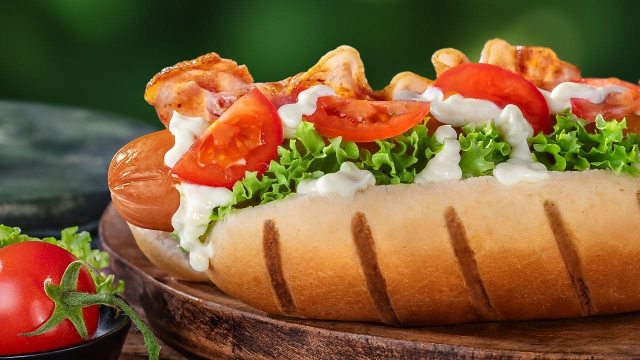 Foto hot dogu Anglie se salátem, rajčetem, majonézou a párkem se slaninou