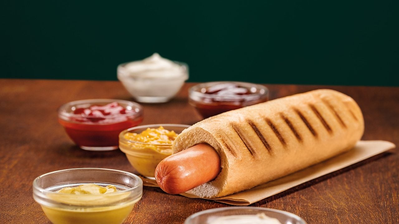 Foto hot dogu Klasik, na pozadí lze vidět hořčici a kečup ve skleněných mističkách
